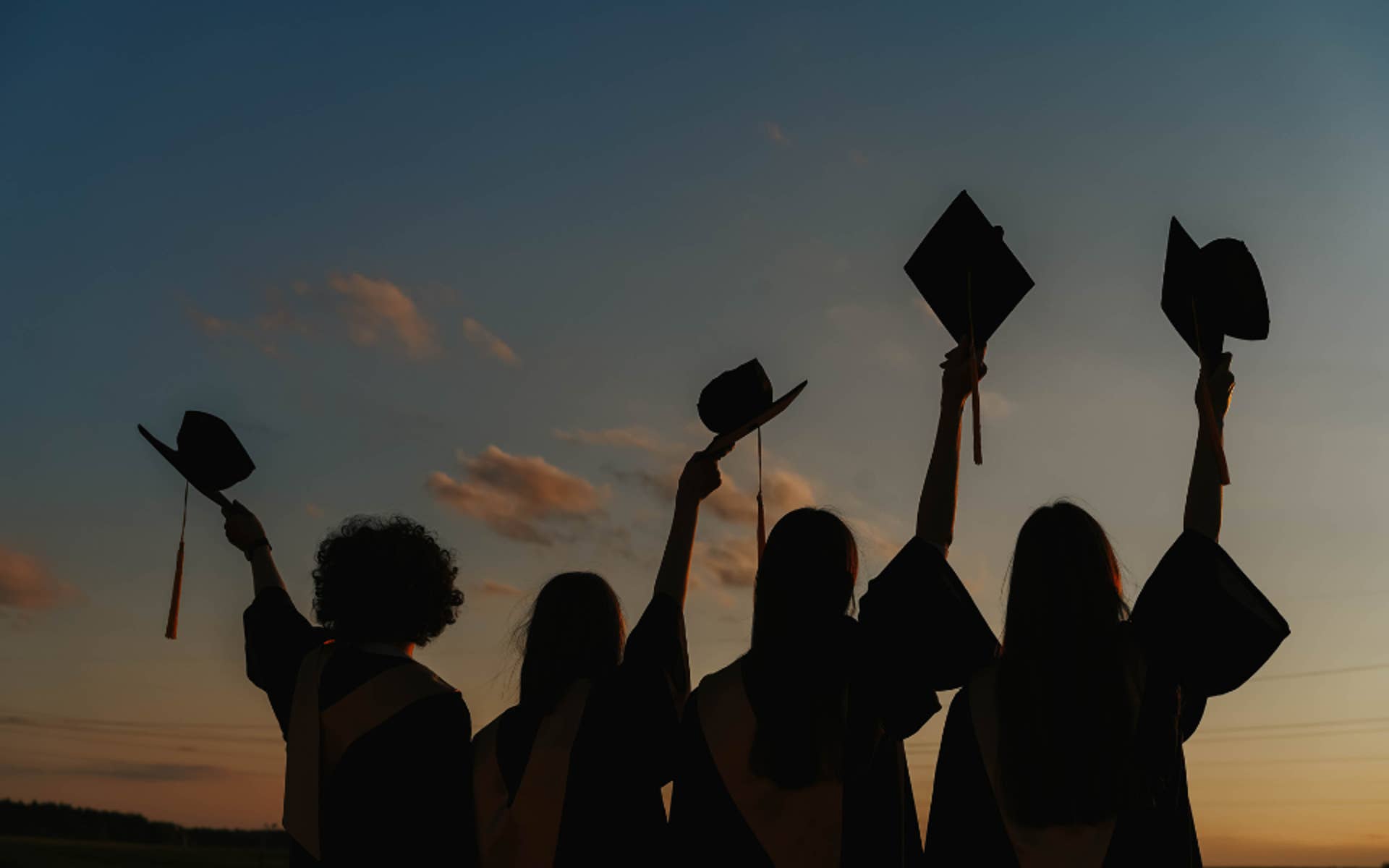 4 youth silhouettes holding graduation caps up in air