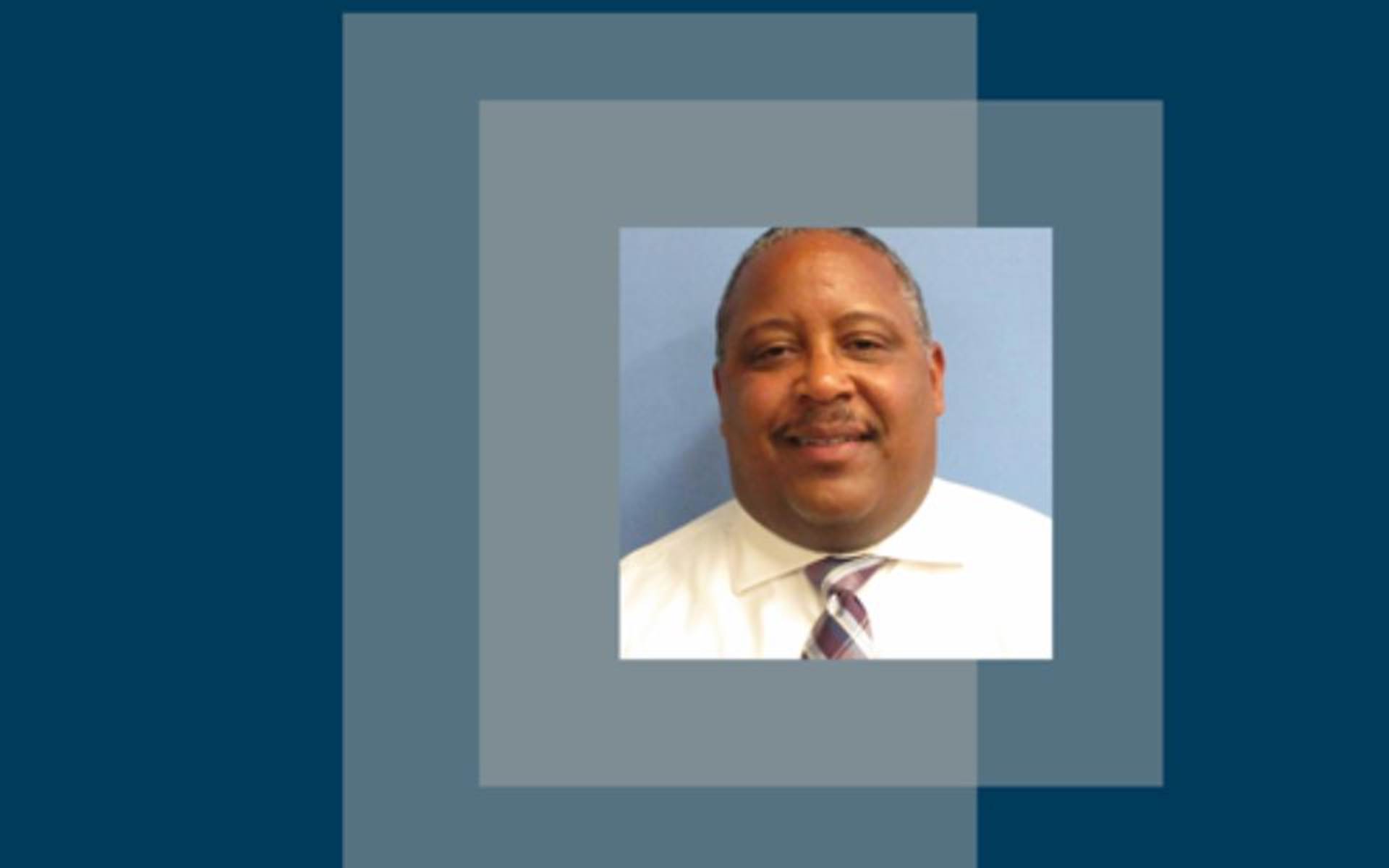 Blue background and a picture of Troy Wharton smiling in a white shirt and tie