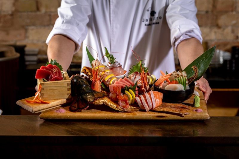 A tray piled high a colourful variety of sushi, sashimi, and maki roll pieces. a chef stands behind, his face cropped out of the photo.