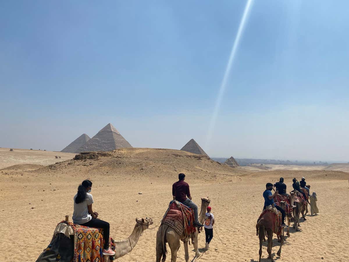 Riding camels toward the pyramids at Giza with beams of light hitting the tips