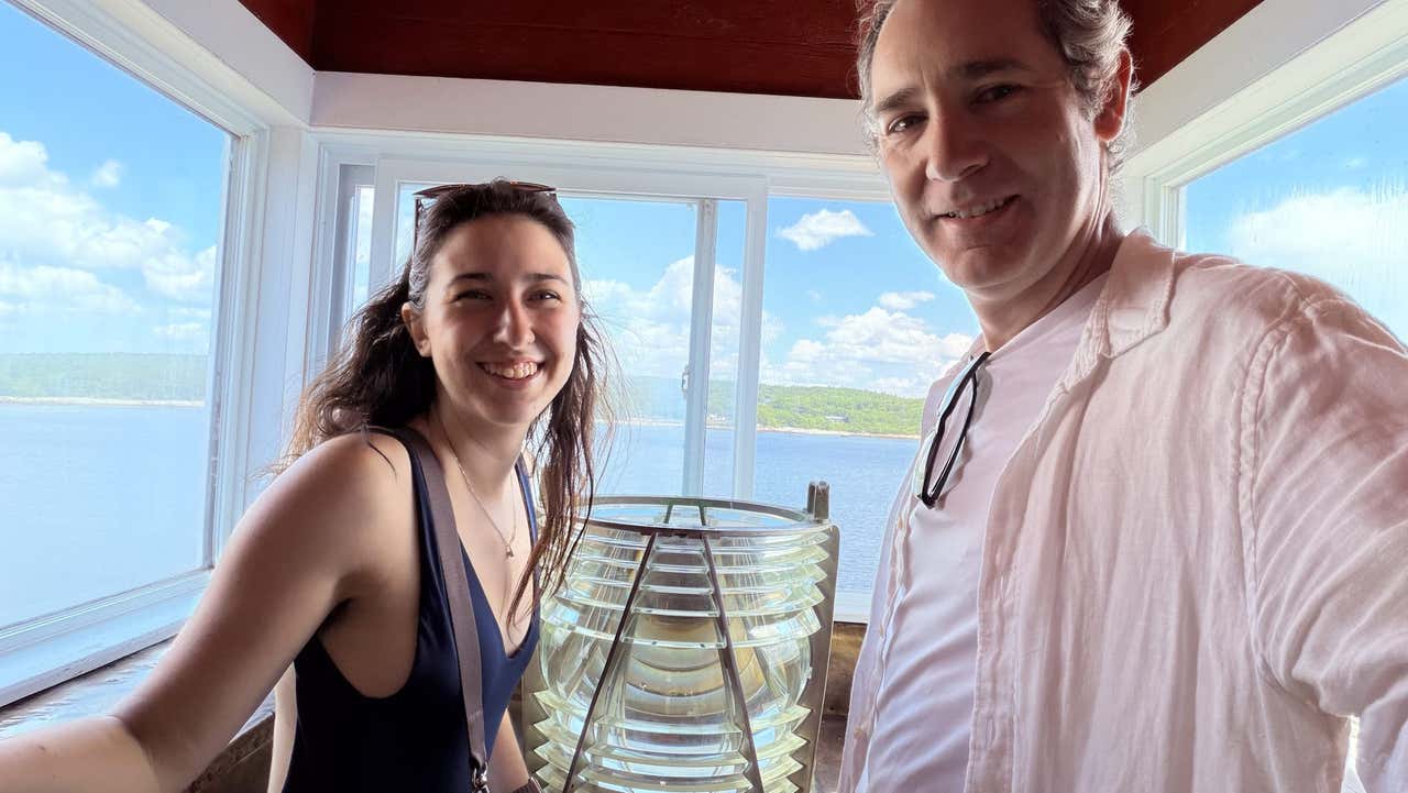 Joel and Gemma smiling in a lighthouse lantern room with the Fresnel lens and the water behind them
