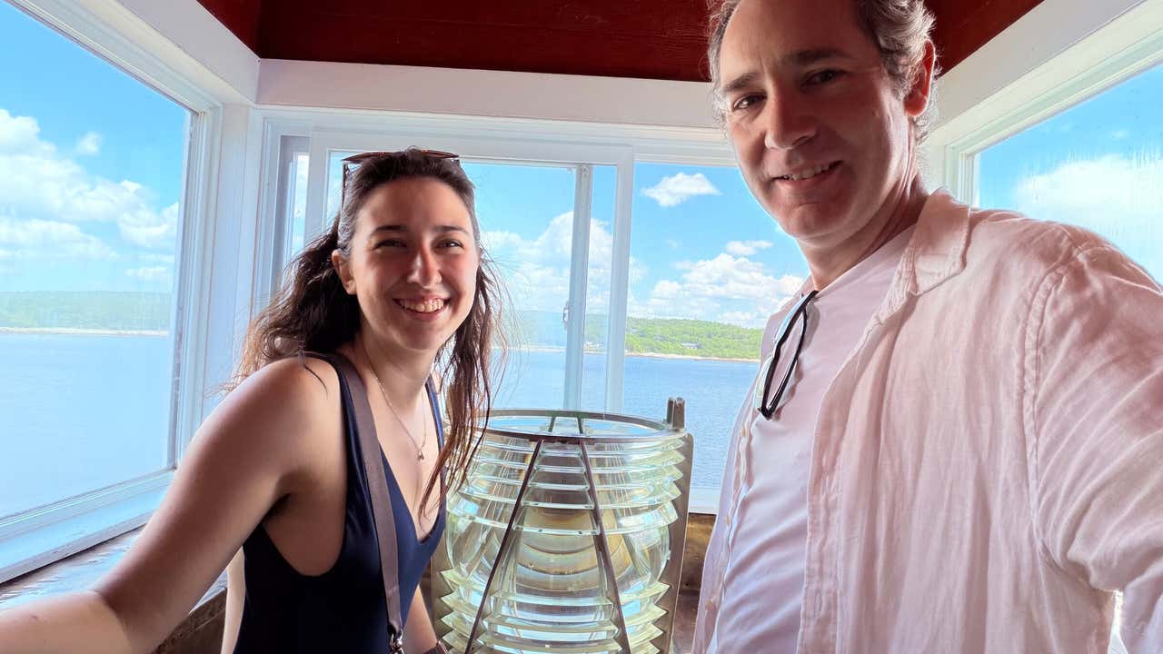 Joel and Gemma smiling inside the Fort Point Lighthouse in Liverpool, water and coastline visible through the windows behind them