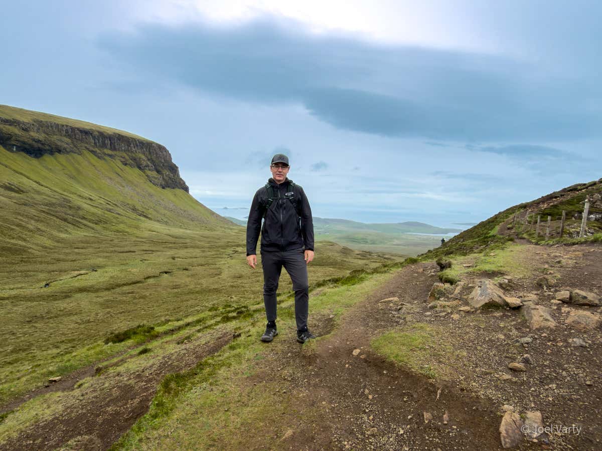 Me on the trail with the great flat wall of the Quiraing behind me