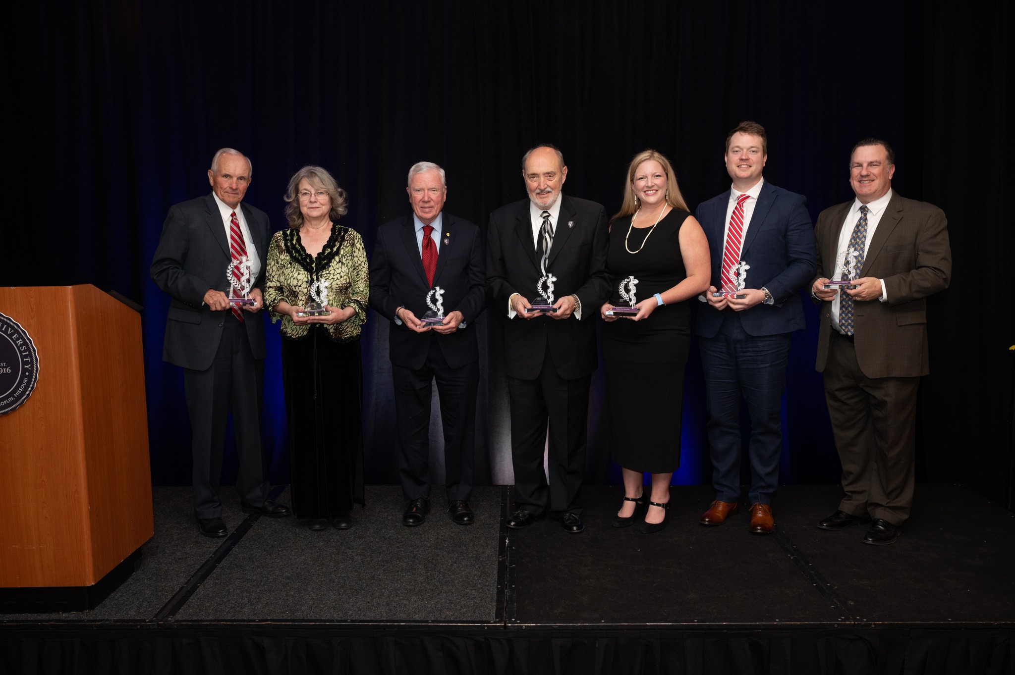 Seven KCU Alumni Award Winners stand together, each holding a trophy in the shape of the caduceus. 