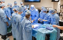 Students learn together around surgical table while in surgical gowns. 