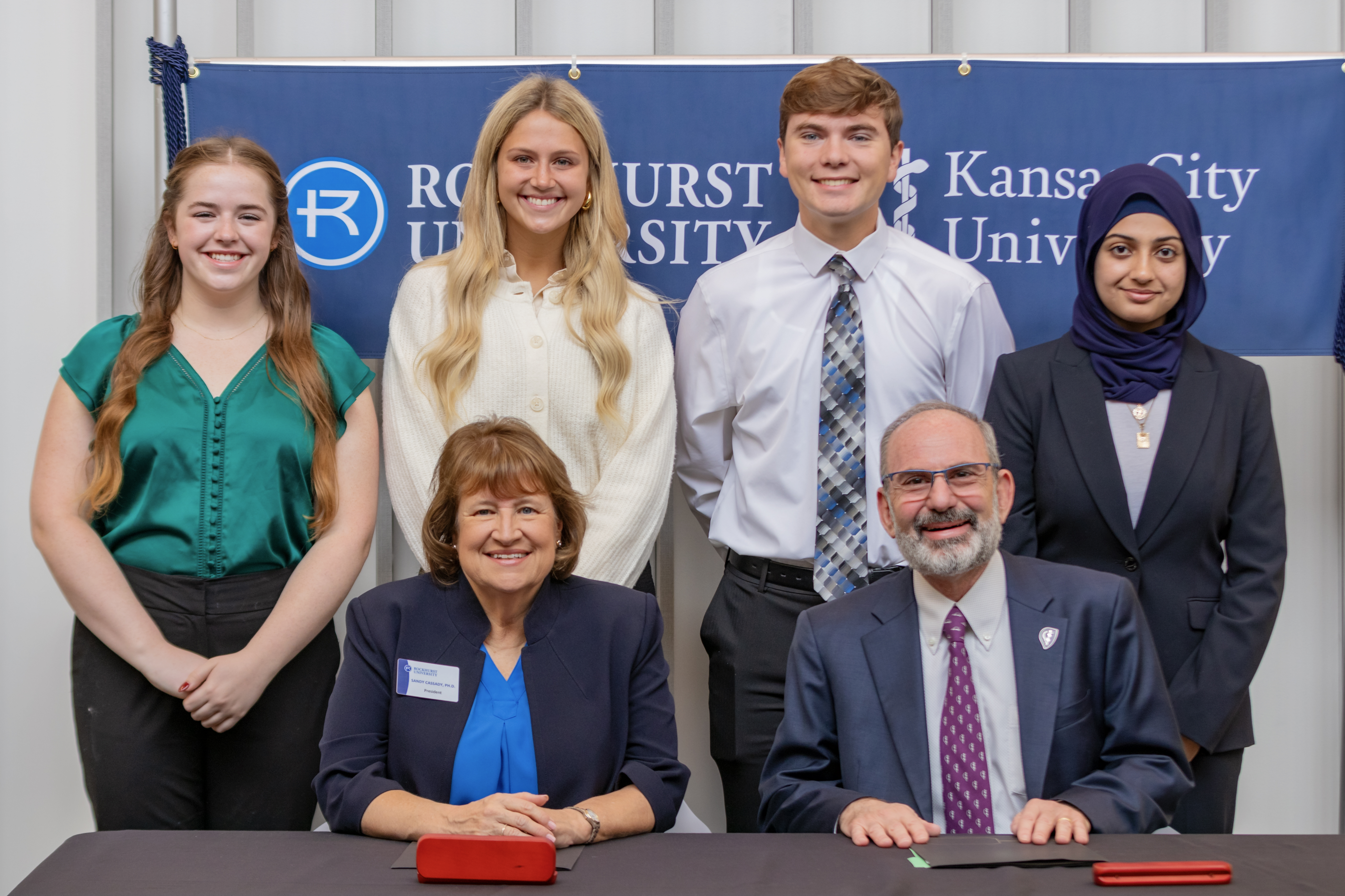 Four Rockhurst University students smile as they stand behind KCU and Rockhurst leaders as they sign agreement. 