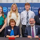 Four Rockhurst University students smile as they stand behind KCU and Rockhurst leaders as they sign agreement. 