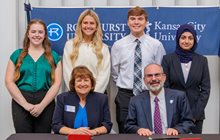 Four Rockhurst University students smile as they stand behind KCU and Rockhurst leaders as they sign agreement. 