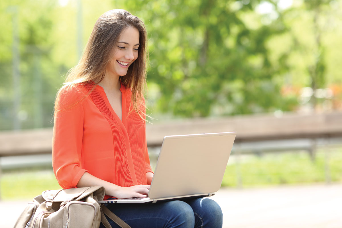 Woman in orange blouse sits on bench outside with laptop in her lap. 