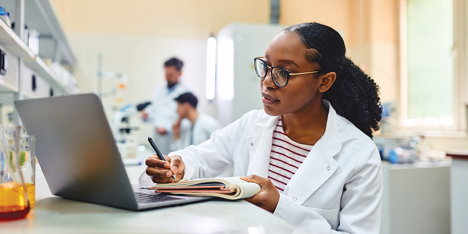 Woman in white coat writes on notepad as she reads from a laptop. 