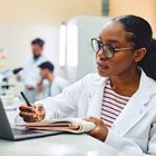 Woman in white coat writes on notepad as she reads from a laptop. 