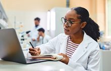 Woman in white coat writes on notepad as she reads from a laptop. 