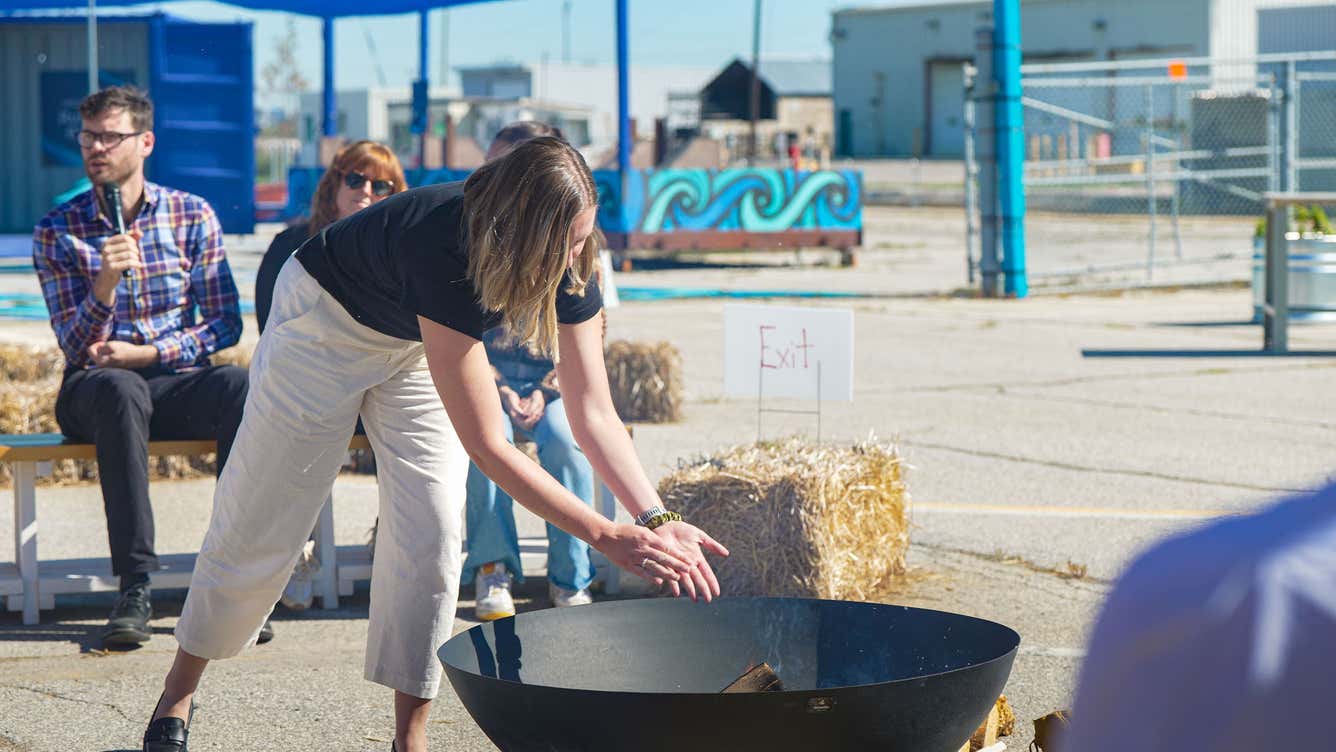 A woman stirs a large black pot over an open fire, cooking a meal outdoors.