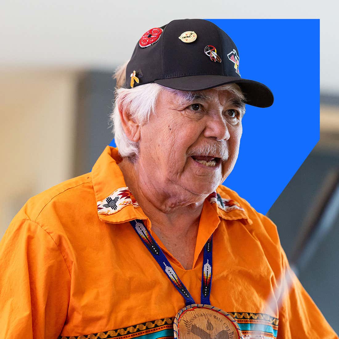 An older man wearing an orange shirt and a hat, smiling while standing outdoors.
