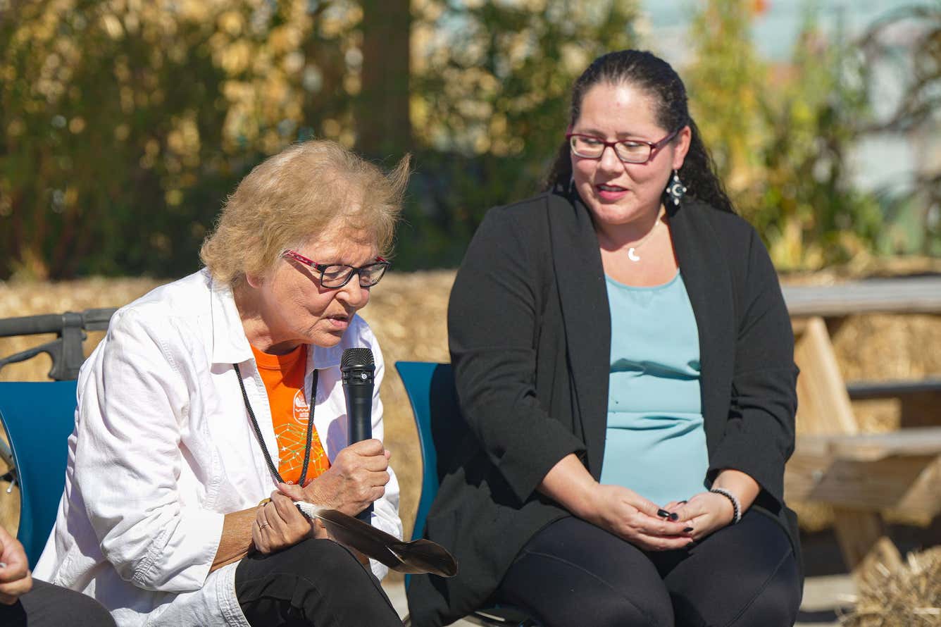A woman seated on a bench, holding a microphone, appears engaged in conversation or an interview.